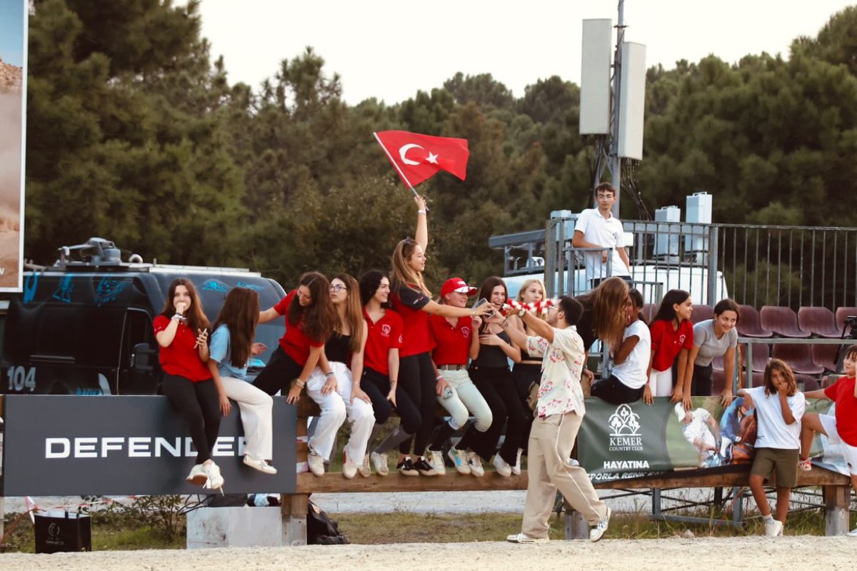 Supporters in red sitting on a barrier at a show jumping competition, one waving a Turkish flag
